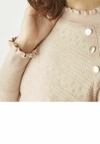 Ljusrosa stickad tröja med strukturerad mönstring, volangkrage och knappdetaljer på framsidan. Nätbild av axeln och ärmen.