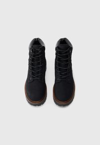 Par de botas de invierno negras con cordones, con cuellos acolchados y suelas intermedias marrones sobre un fondo blanco liso.