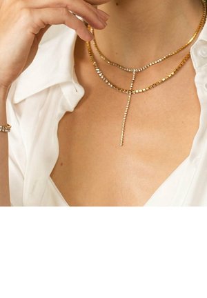 Personne portant des colliers superposés en or et argent ainsi qu'une chemise blanche au col ouvert, main près du cou.