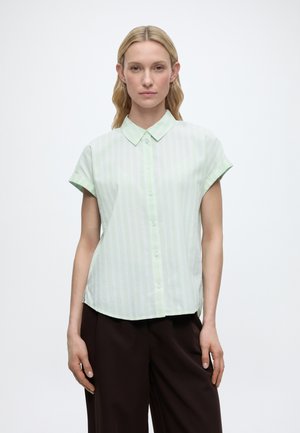 Blondynka z rozpuszczonymi włosami, ubrana w krótkim rękawem miętową, paskowaną koszulę zapinaną na guziki oraz ciemnobrązowe spodnie, stojąca na białym tle.