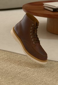 Stivale in pelle marrone con lacci e suola bianca che fluttua sopra un tappeto beige testurizzato, vicino a un tavolo rotondo in legno con libri impilati.