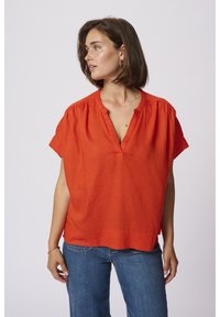 Blouse en lin rouge avec un décolleté en V, manches courtes et détails froncés aux épaules, associée à un jean bleu. Texture lisse, coupe décontractée.