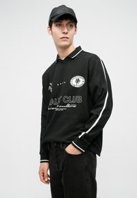 Giovane uomo che indossa un berretto nero e una maglietta a maniche lunghe nera con strisce bianche e testo grafico, mentre posa con una mano in tasca.