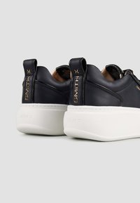Sneaker in pelle nera con suola piatta bianca, presentano dettagli dorati e branding sul tallone, texture liscia e forma arrotondata.
