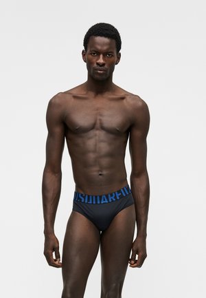 Homme musclé portant un slip noir avec une ceinture bleue marquée, debout face à un fond clair uni.