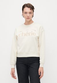 Kremfarget genser med cropped design, ribbestrikket kant og mansjetter, med ordet "chérie" i en lys grafisk trykk. Mørke jeans bæres under.