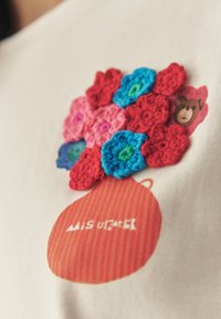 Biela tričko s farebným dizajnom kvetinovej kytice, na ktorom sú červené, modré, ružové a zelené háčkované kvety a červený hrniec s bielym textom.