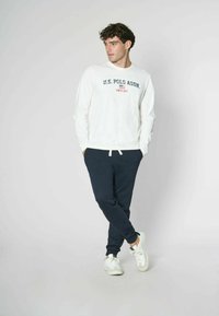 Maglietta a maniche lunghe bianca con la scritta "U.S. Polo Assn." stampata in blu scuro e rosso. Pantaloni joggers blu navy con vita elasticizzata e sneakers bianche.