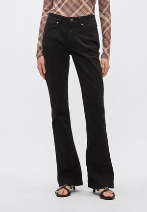 Jean noir évasé avec une texture lisse, des coutures discrètes, des poches avant et une fermeture à bouton. Associé à des sandales noires à brides.