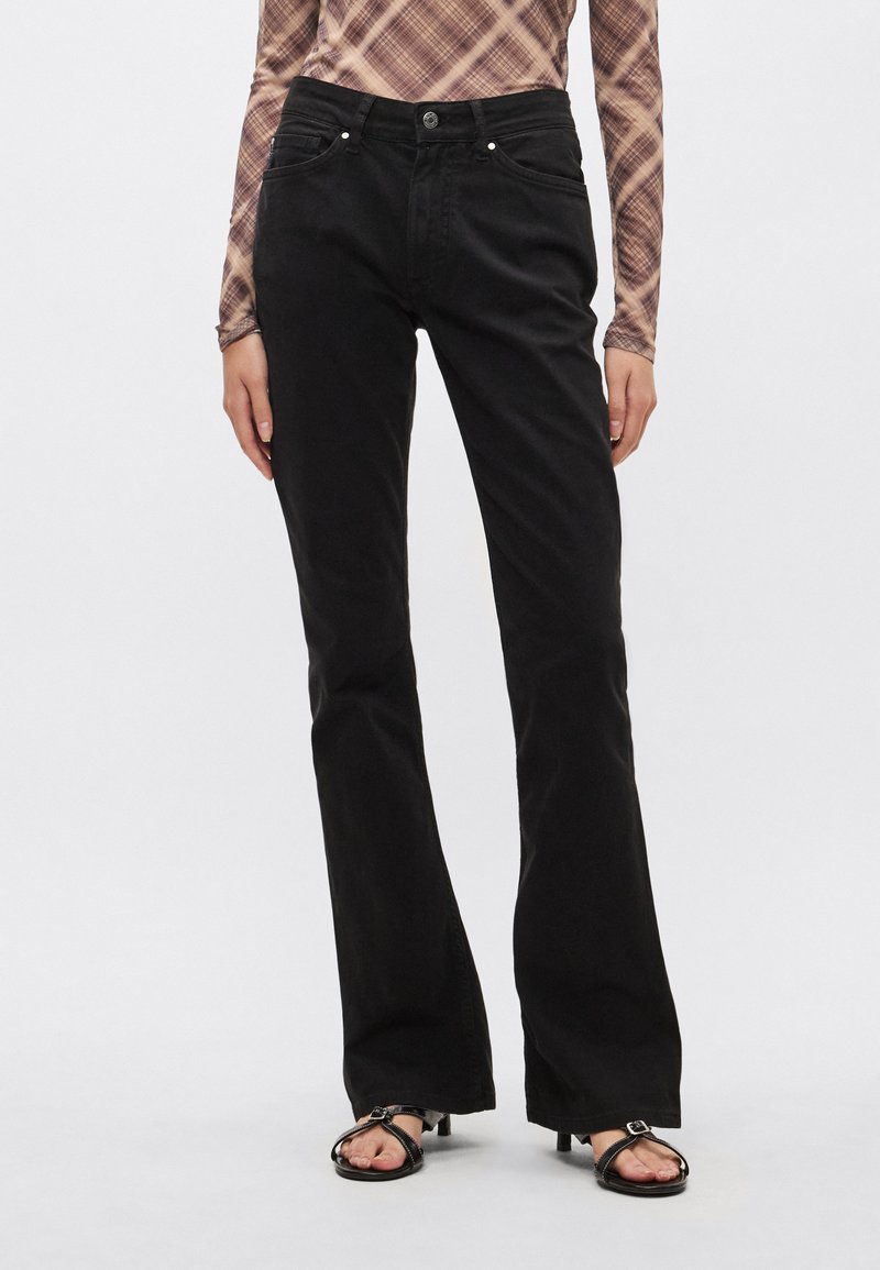 Zwarte jeans met uitlopende pijpen en een gladde textuur, subtiele stiksels, voorzakken en een knoopsluiting. Gecombineerd met zwarte sandalen met bandjes.