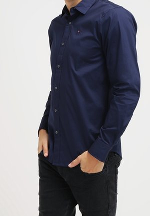 Mann trägt ein tailliertes dunkelblaues Hemd mit Knöpfen und langen Ärmeln, steht mit den Händen in den Taschen einer schwarzen Jeans vor einem schlichten Hintergrund.