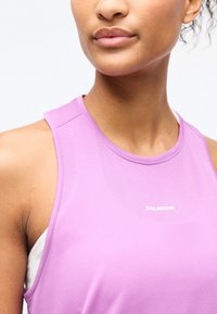 Κοντινό πλάνο ενός ατόμου που φοράει αθλητικό τοπ χωρίς μανίκια σε λιλά χρώμα με το λογότυπο Salomon στο στήθος.