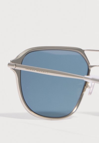 Lunettes de soleil avec monture en métal argenté, verres teintés bleus et branches fines, montrant partiellement la gravure "ISABEL MARANT" sur la branche.