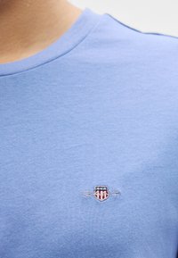 Modra bombažna majica s kroglo izrezom in majhnim vezenim logotipom s ščitnim dizajnom na levi strani prsnega koša. Gladka tekstura.