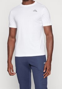 Bílý bavlněný tričko s krátkým rukávem a kulatým výstřihem. Na levé straně hrudi se nachází černé logo Kappa. Hladká textilie.