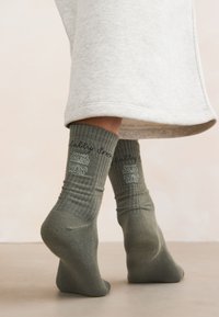 Pieds portant des chaussettes vertes côtel ées avec des motifs brodés, se tenant sur la pointe des pieds, partiellement couverts par un vêtement gris clair.