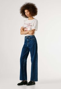 Bela majica s tiskanim vzorcem, združena z visokim pasom, širokimi temi modrimi kavbojkami in črnimi čevlji. Gladek material in sproščen kroj.