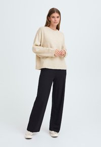 Pull en tricot beige avec un col rond, des poignets et un ourlet côtelés. Associé à un pantalon noir à jambes larges taille haute et des baskets décontractées.