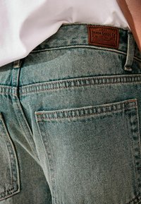 Jean en denim bleu clair avec un tissage texturé, deux poches arrière et une étiquette en cuir marron sur la taille.