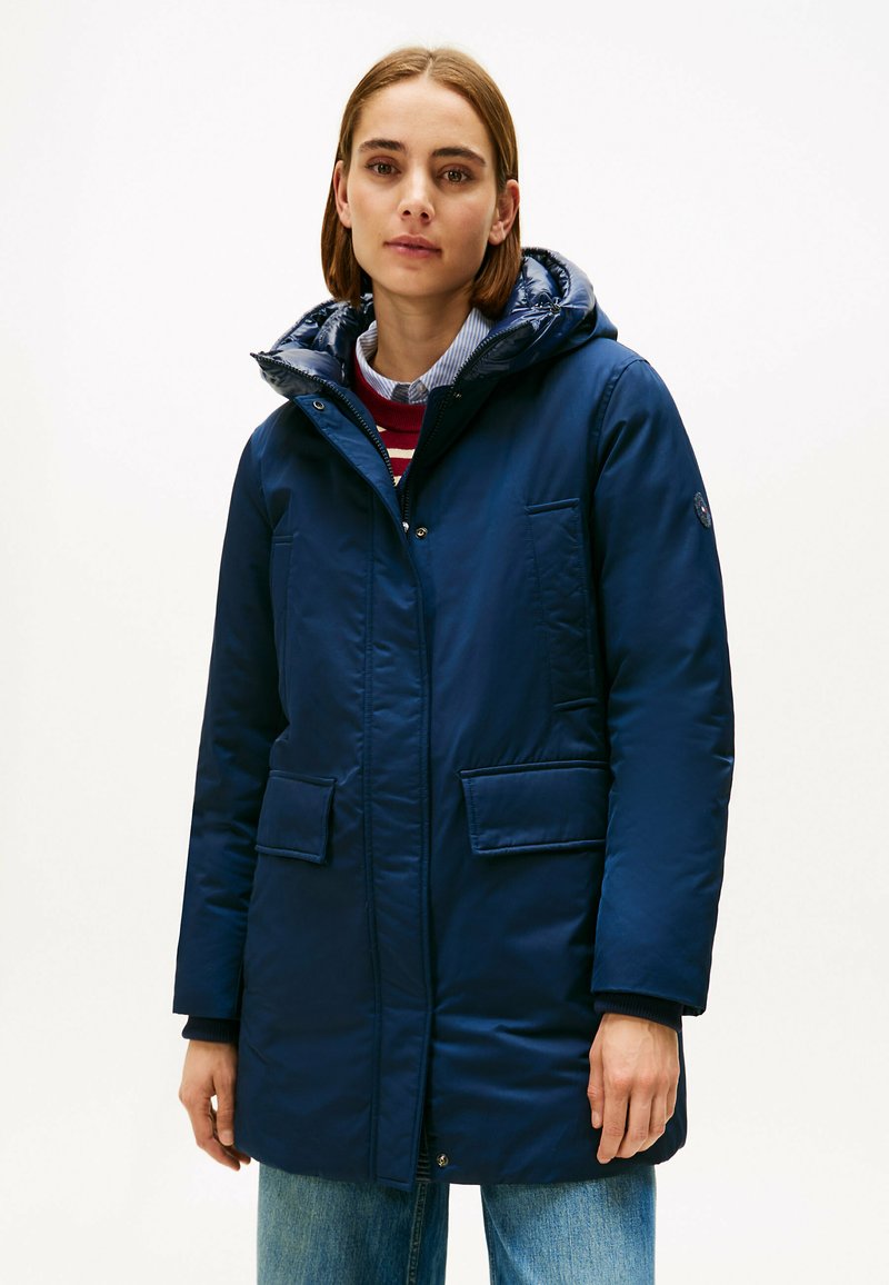 Giacca isolante blu navy con cappuccio, dotata di chiusura a zip, due tasche frontali e una texture liscia. Design casual con una vestibilità leggermente sagomata.