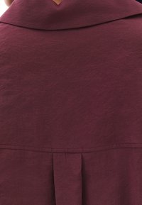 Indumento maroon con un tessuto texturizzato. Presenta un ampio colletto e dettagli di pieghe verticali sul retro, che esaltano il design minimalista.