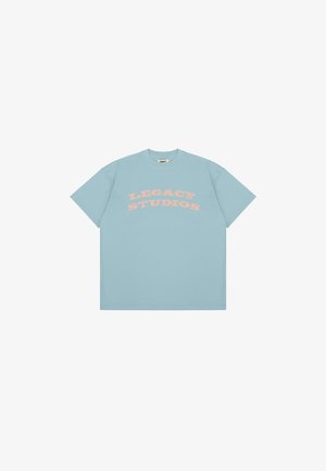 Šviesiai mėlynas medvilninis trumpas rankovėmis marškinėliai su ryškiu, kreiviu tekstu rožinės spalvos: "LEGACY STUDIOS." Paprastas dizainas su laisvu siluetu.