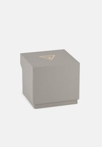 Boîte carrée texturée grise avec un triangle doré du logo Guess sur le dessus, placée sur fond blanc.