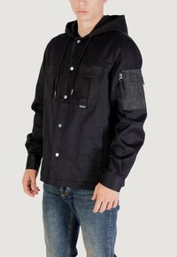 Chaqueta negra con capucha, cierre frontal con botones, dos bolsillos en el pecho y un bolsillo con cremallera en la manga. Fabricada en una tela suave con un corte relajado.