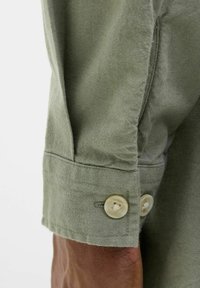 Puño de camisa de color verde oliva con dos botones crema, con una tela suave y texturizada y un detalle de costura limpio.