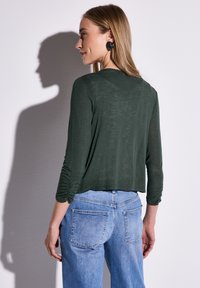 Grön, långärmad topp med en avslappnad passform och rynkade detaljer på ärmarna, ihop med ljusblå, löst sittande jeans.