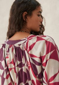 Blouse à manches courtes avec des motifs géométriques violets et rouges, présentant un gathering au niveau du cou à l'arrière et un tissu doux et texturé.
