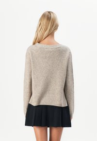 Mujer rubia vestida con un suéter de punto beige y una falda plisada negra, vista desde la espalda contra un fondo blanco.