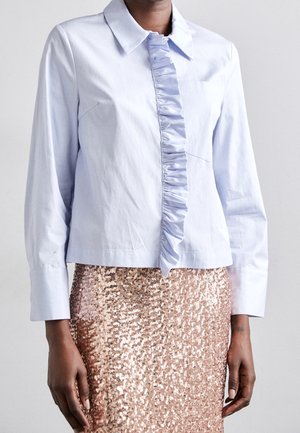 Woman wearing a light blue long-sleeve shirt with ruffled front detail paired with a rose gold sequined skirt.
