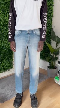 Persona vestida con jeans de mezclilla azul claro desgastados, mocasines negros con borlas y una camiseta blanca de manga larga con mangas negras que presentan texto gótico blanco.