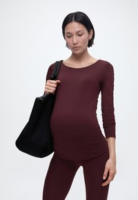 Rase naine burgundia pikkade varrukatega pluusis ja retuusides hoiab suurt musta õlakotti, vaadates alla neutraalse ilmega.