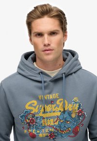 Sudadera de algodón azul con un estampado gráfico que presenta motivos florales y de dragón, cuello con cordón y puños acanalados.