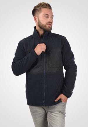 Navy fleece jacket featuring a black panel and zippered chest pocket, high collar, and full front zipper. Soft texture, casual fit.
