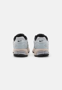 Sapatilhas Nike com superior de malha cinza, detalhes em preto, colarinho acolchoado e uma sola texturizada bege e cinza. A vista traseira exibe o detalhe do logotipo.