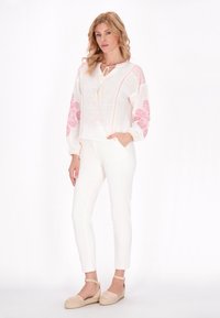 Weiße Leinenbluse mit rosa Blumenstickerei, ausgestattet mit einem gebundenen Ausschnitt und langen Ärmeln, kombiniert mit weißen, eng anliegenden Hosen und beigen Espadrilles.
