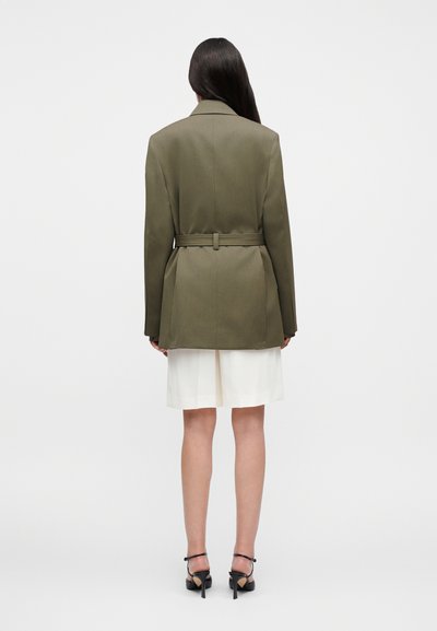 Γυναίκα με μακριά σκούρα μαλλιά που φοράει πράσινο μπλέιζερ με ζώνη, λευκή φούστα και μαύρα σανδάλια με ψηλό τακούνι, στέκεται απέναντι σε λευκό φόντο.