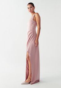 Rožinė satino suknelė su giliu šonu plyšiu, prigludusia bodice ir plisuotomis detalėmis. Modelis vilki blizgančius smailius batelius ir auskarus.