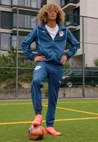 Chándal deportivo azul hecho de material ligero, con una chaqueta con cremallera y un logo, combinado con botas de fútbol rosa brillante sobre un balón.
