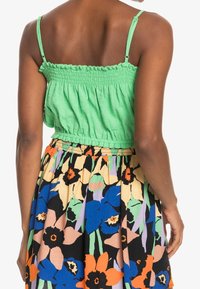 Vihreä smokkikuvioinen lyhythihainen toppi, joka on yhdistetty kukkakuosiseksi hameeksi, jossa on eloisia värejä kuten oranssia, sinistä ja vihreää.