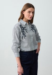Gestreiftes Hemd in Weiß und Marineblau mit floraler Stickerei auf der Vorderseite, verfügt über lange Ärmel und einen Kragen. Kombiniert mit dunklen Jeans.