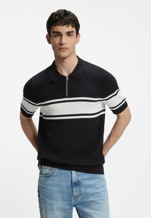 Junger Mann mit kurzen dunklen Haaren, der ein schwarz-weiß gestreiftes kurzärmeliges Strick-Poloshirt und hellblaue Jeans trägt, steht vor weißem Hintergrund.
