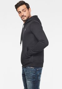 Svart huvtröja med framficka, dragband och logodetalj. Bärs med blå jeans med ljus tvätt och blekt utseende.