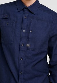Marineblauwe button-up shirt van textuurstof, met een kleine patchzak, zwarte knopen en een merketiket op de borst.