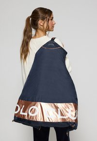 Gilet rembourré bleu marine avec une bande bronze brillante à l'ourlet, comportant un texte de marque en relief. Texture lisse, léger et avec une fermeture éclair ajustable.