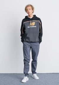Melnas kapuces džemperis ar oranžiem akcentiem, kurā ir "New Balance" logo, kombinēts ar pelēkiem biksēm un baltām kedām. Modelis stāv ar rokām kabatās.