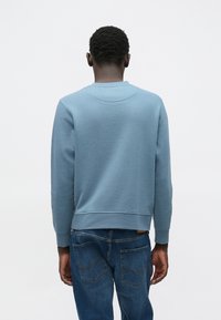 Gaiši zils džemperis ar garām piedurknēm, apaļu kaklu un mīkstu tekstūru; valkāts kopā ar tumši ziliem džinsiem, kam aizmugurē šuves un brīvs piegriezums.